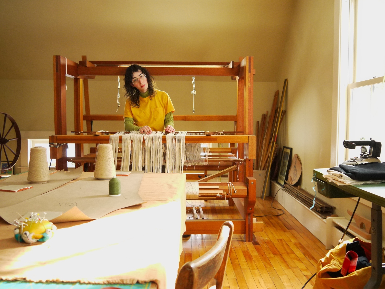 a white trans woman stands inside the wooden skeleton of a loom. in the middle of working the threads, she looks up at the camera. her curly hair is still drying. the studio is full of winter light and warm wood.